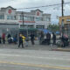People line up along a sidewalk beside a chain-link fence in front of a building, with bags and belongings scattered nearby.