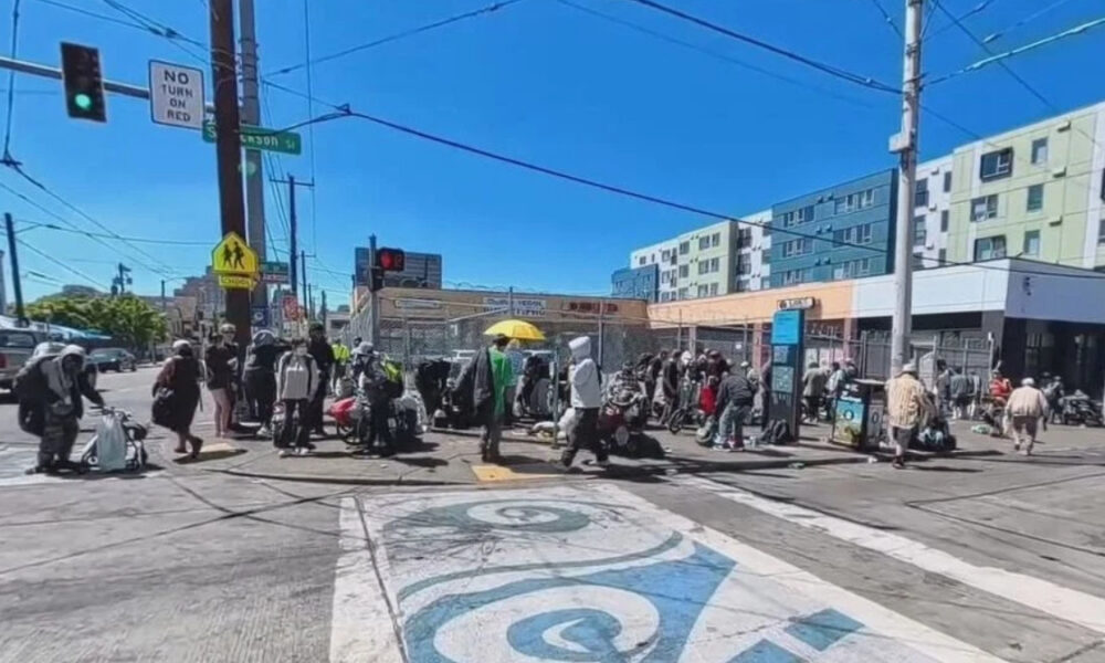 Crowd of pedestrians carrying bags and luggage crossing a busy urban intersection on a sunny day with blue sky.