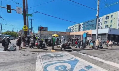 Crowd of pedestrians carrying bags and luggage crossing a busy urban intersection on a sunny day with blue sky.