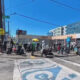 Crowd of pedestrians carrying bags and luggage crossing a busy urban intersection on a sunny day with blue sky.