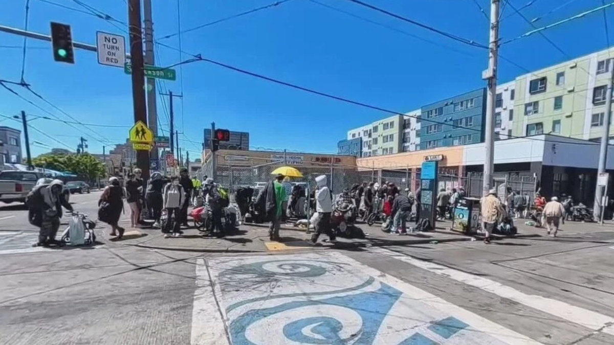 Crowd of pedestrians carrying bags and luggage crossing a busy urban intersection on a sunny day with blue sky.