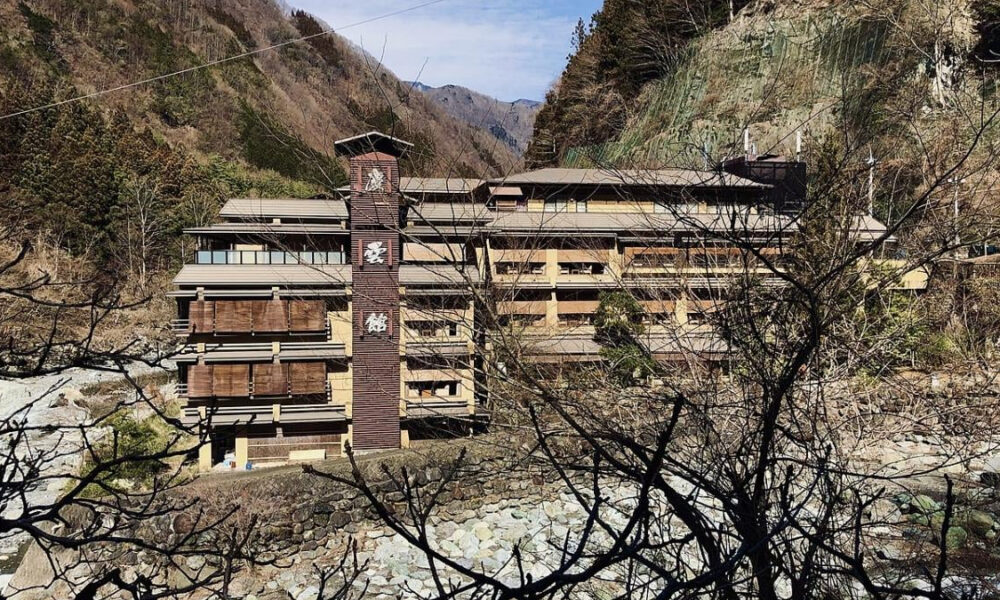 Mountain resort-style wooden lodge with a tall central tower, set in a rocky valley; bare branches frame the foreground.