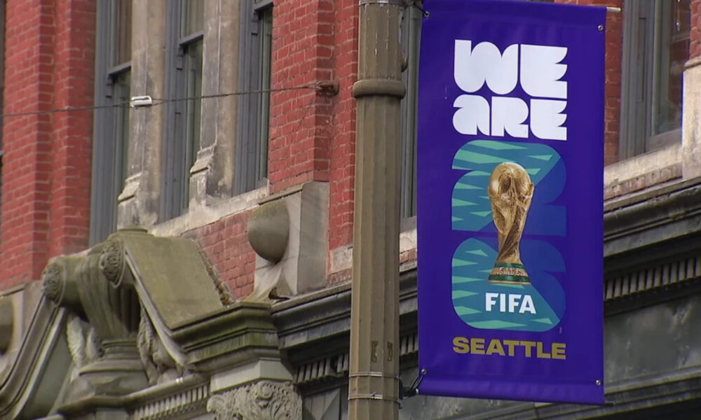 Purple street banner reading 'WE ARE' with the FIFA World Cup trophy and the word 'SEATTLE' at the bottom, hung on a lamppost.