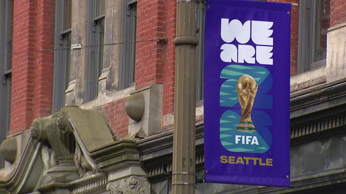 Purple street banner reading 'WE ARE' with the FIFA World Cup trophy and the word 'SEATTLE' at the bottom, hung on a lamppost.