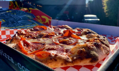 Pepperoni pizza in a box on a red-and-white checkered liner outdoors, sunlight on cheese and pepperoni slices.