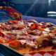Pepperoni pizza in a box on a red-and-white checkered liner outdoors, sunlight on cheese and pepperoni slices.