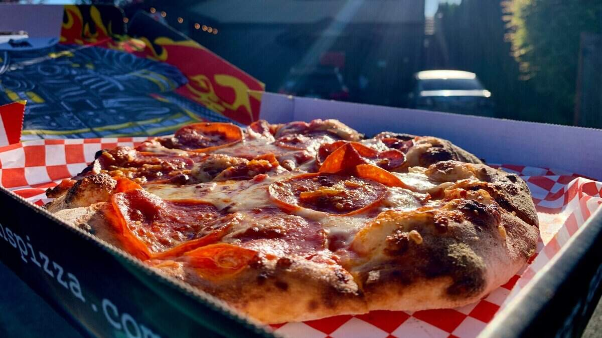 Pepperoni pizza in a box on a red-and-white checkered liner outdoors, sunlight on cheese and pepperoni slices.
