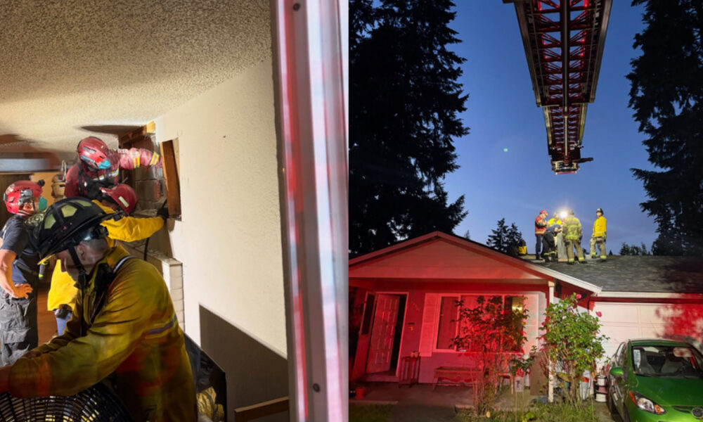 Firefighters breach a wall inside a house while others stand on a roof with a ladder during a nighttime emergency travel scene.