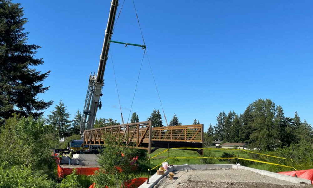 Crane lifts a wooden pedestrian bridge into place at a construction site under a clear blue sky, with yellow caution tape in the background.