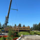 Crane lifts a wooden pedestrian bridge into place at a construction site under a clear blue sky, with yellow caution tape in the background.