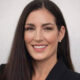 Professional headshot of a smiling woman with long dark hair wearing a black blazer and pearls against a neutral background.
