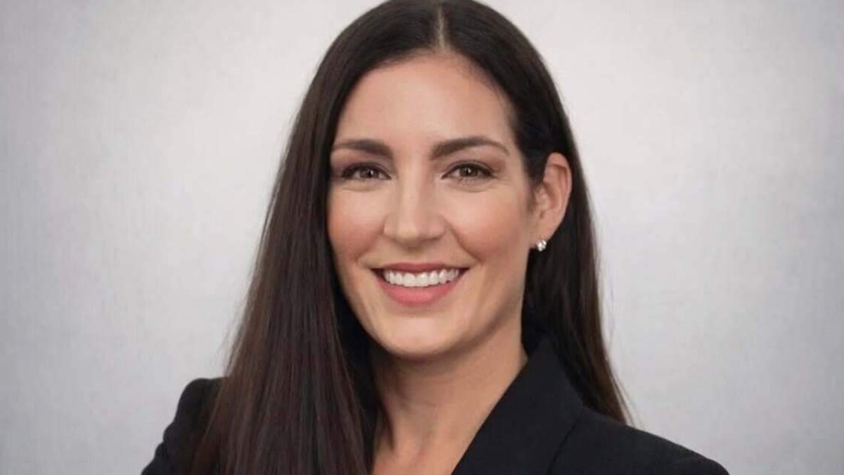 Professional headshot of a smiling woman with long dark hair wearing a black blazer and pearls against a neutral background.