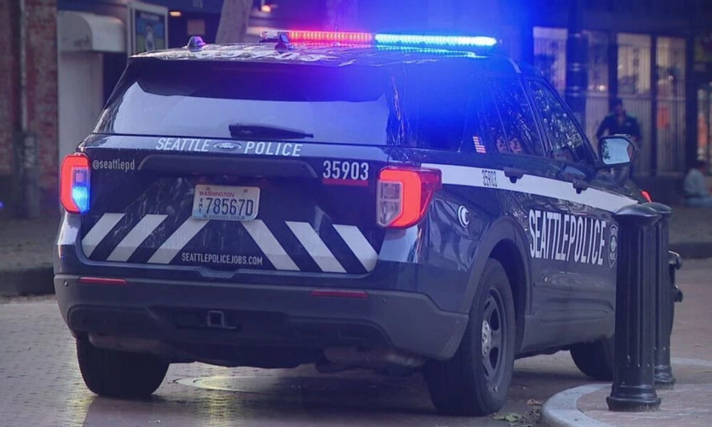 Seattle Police SUV with flashing red and blue lights parked on a city street, rear view shows 'SEATTLE POLICE' lettering and striped rear design.
