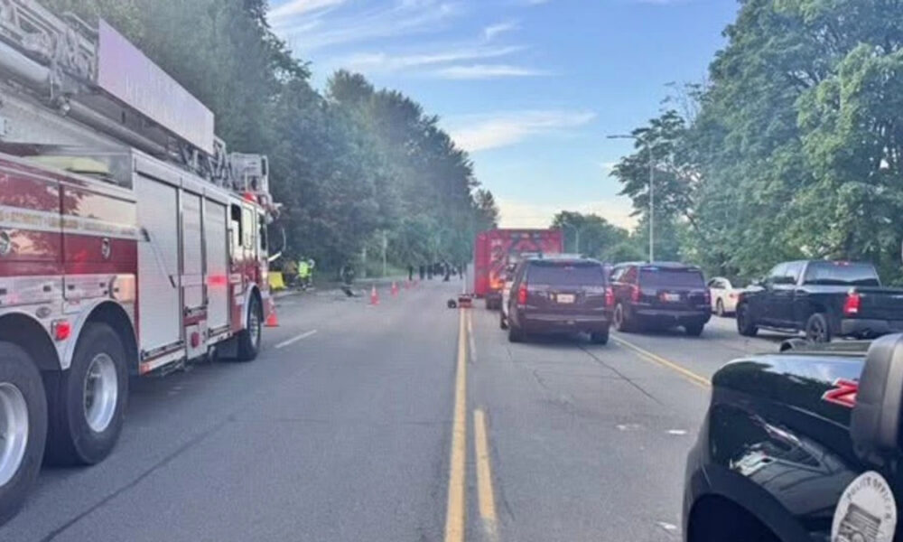 Fire truck blocks a two-lane road as firefighters work near a crash; cones mark the scene with cars stopped on both sides under a blue sky.