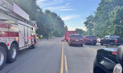 Fire truck blocks a two-lane road as firefighters work near a crash; cones mark the scene with cars stopped on both sides under a blue sky.