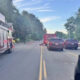 Fire truck blocks a two-lane road as firefighters work near a crash; cones mark the scene with cars stopped on both sides under a blue sky.