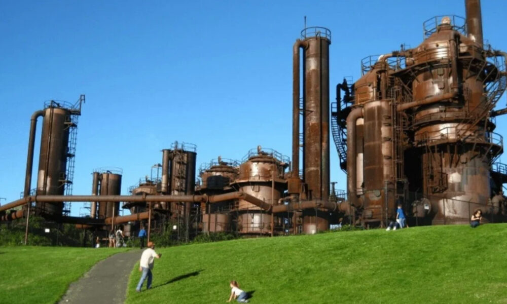 Rusty industrial refinery with tall towers and interconnected pipes against a bright blue sky, people on a grassy hill in the foreground.