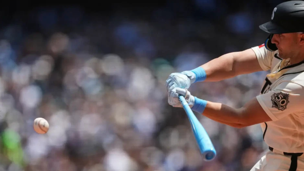 Baseball batter in a white uniform swings a blue bat as a pitched ball approaches, with a blurred crowd in the background.