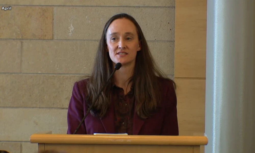 Woman in a burgundy blazer speaks at a wooden podium with a microphone in a conference room.