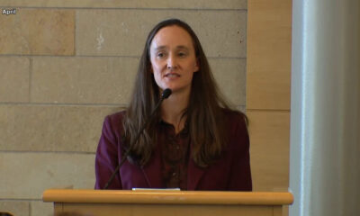 Woman in a burgundy blazer speaks at a wooden podium with a microphone in a conference room.