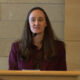 Woman in a burgundy blazer speaks at a wooden podium with a microphone in a conference room.