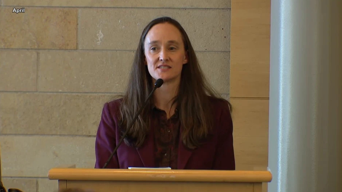 Woman in a burgundy blazer speaks at a wooden podium with a microphone in a conference room.
