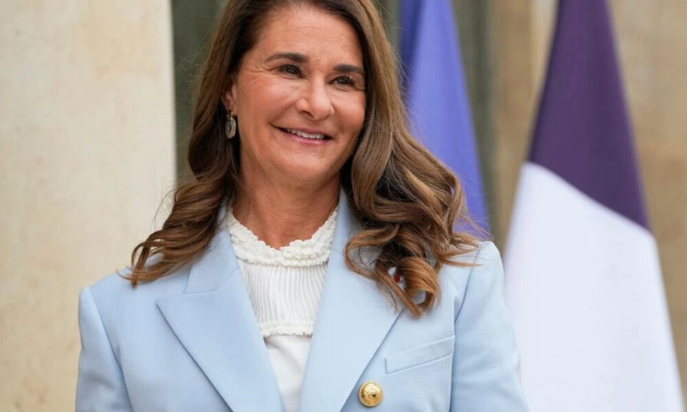 Smiling woman in a light blue blazer outdoors, with flags in the background, posing for a formal photo.