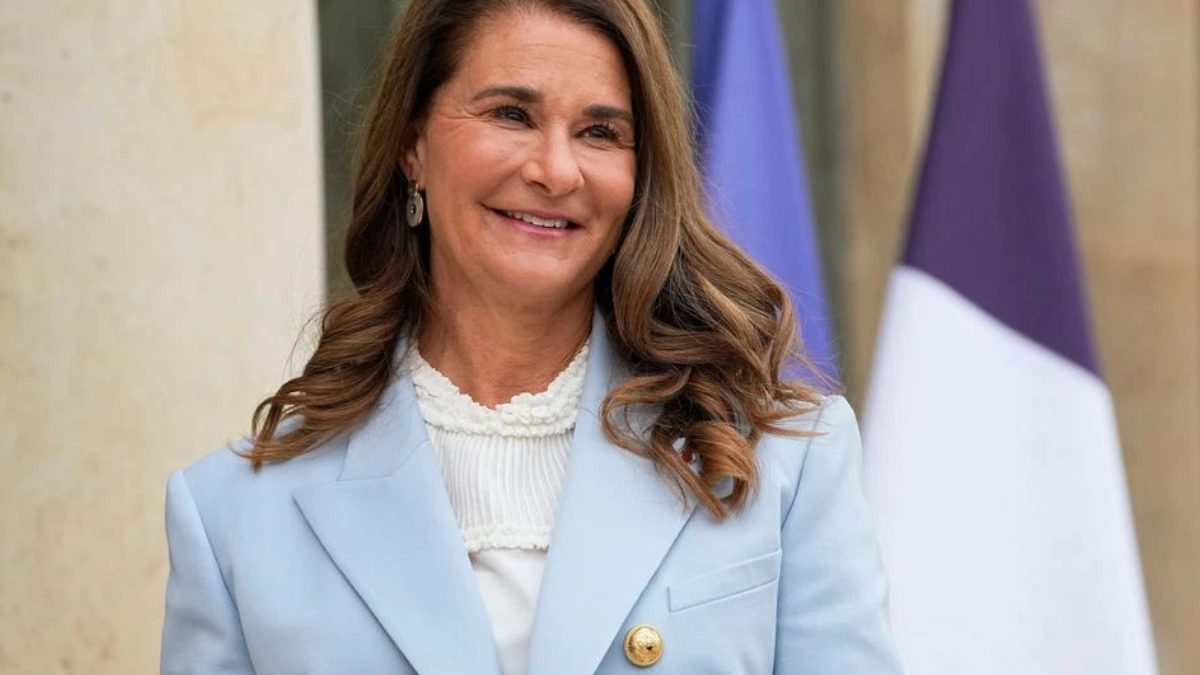 Smiling woman in a light blue blazer outdoors, with flags in the background, posing for a formal photo.