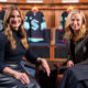 Two women sit in a locker room on orange chairs, smiling, with team jerseys hanging behind them.