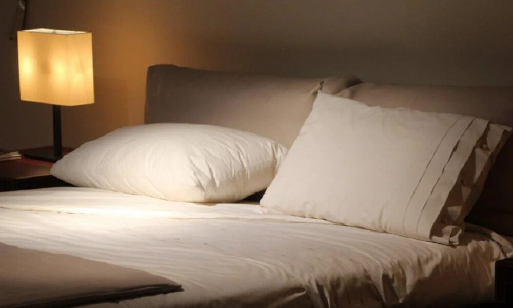 Bed with white sheets and two pillows, beige headboard, warm bedside lamp.