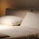 Bed with white sheets and two pillows, beige headboard, warm bedside lamp.