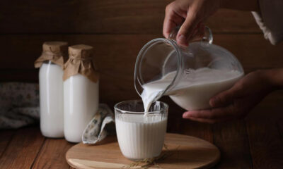Hands pour milk from a glass pitcher into a ribbed glass on a wooden tray; two glass bottles of milk with paper caps in the background.