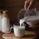 Hands pour milk from a glass pitcher into a ribbed glass on a wooden tray; two glass bottles of milk with paper caps in the background.