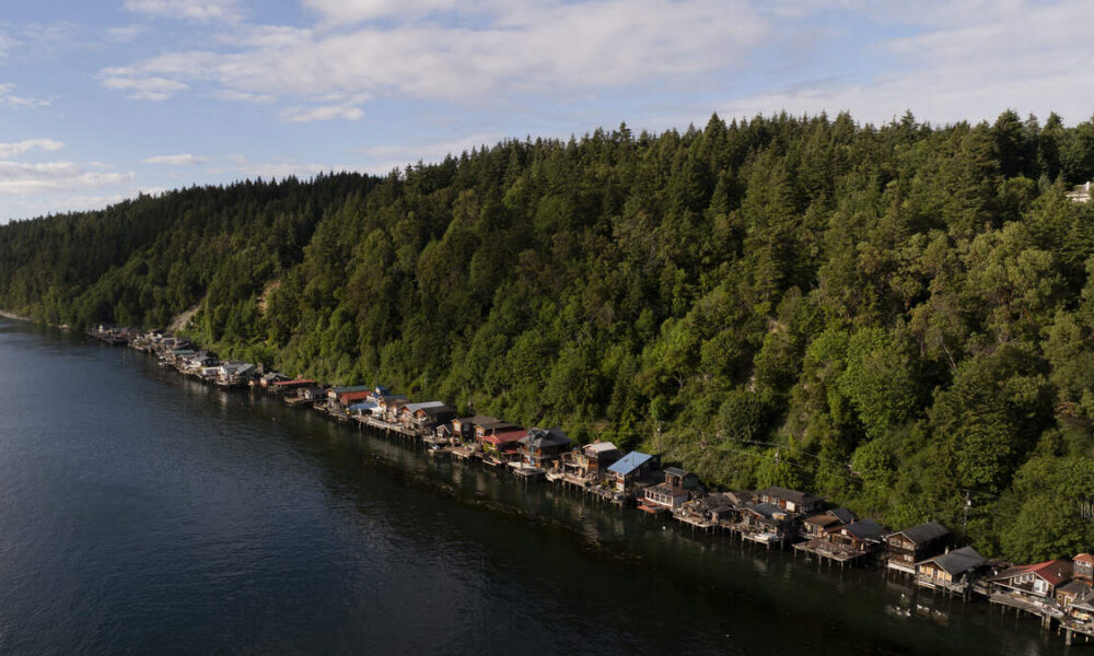 Row of colorful boathouses lining a calm lake with a forested hillside in the background.