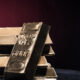Stack of gold bars on a dark reflective surface with a reddish glow in the background, conveying wealth and precious metal storage