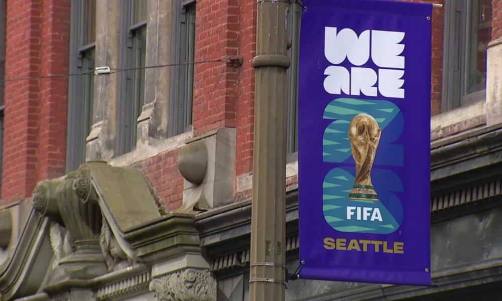 Purple street banner reading 'WE ARE FIFA' with a World Cup trophy image and 'Seattle' at the bottom, hanging on a lamppost by a brick building