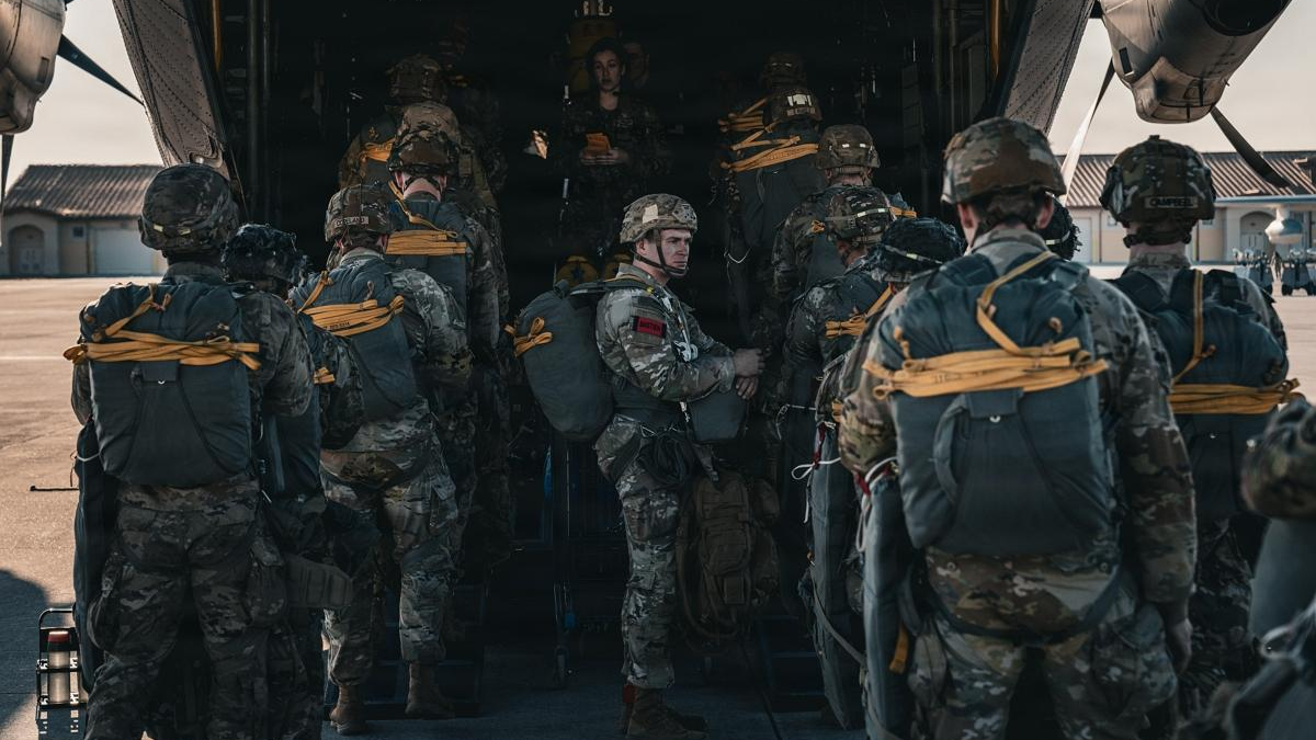 Soldiers in camouflage with backpacks lining up to board a large military transport aircraft via its open rear ramp.