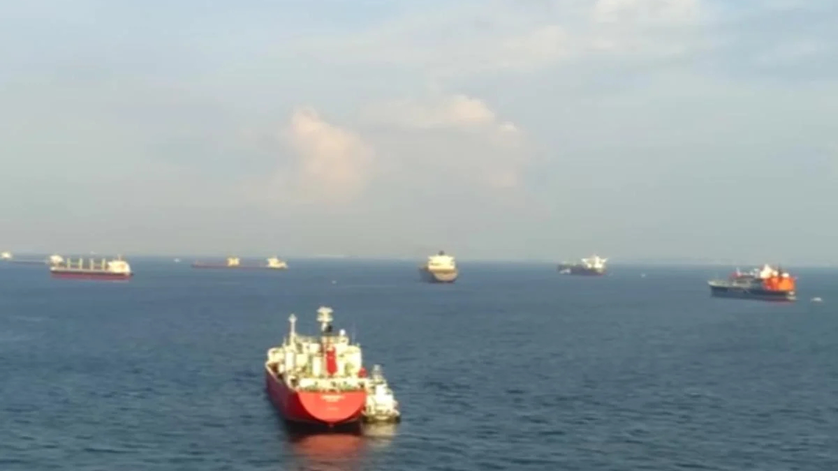 Several ships sail on a calm sea with a red-hulled vessel closest to the foreground and others in the distance under a partly cloudy sky.