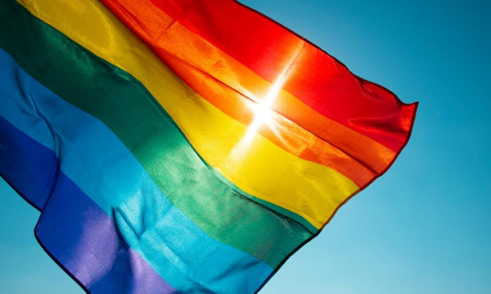 Rainbow Pride flag fluttering in a bright blue sky, sunlight gleaming through the fabric beneath a red stripe.