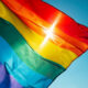 Rainbow Pride flag fluttering in a bright blue sky, sunlight gleaming through the fabric beneath a red stripe.