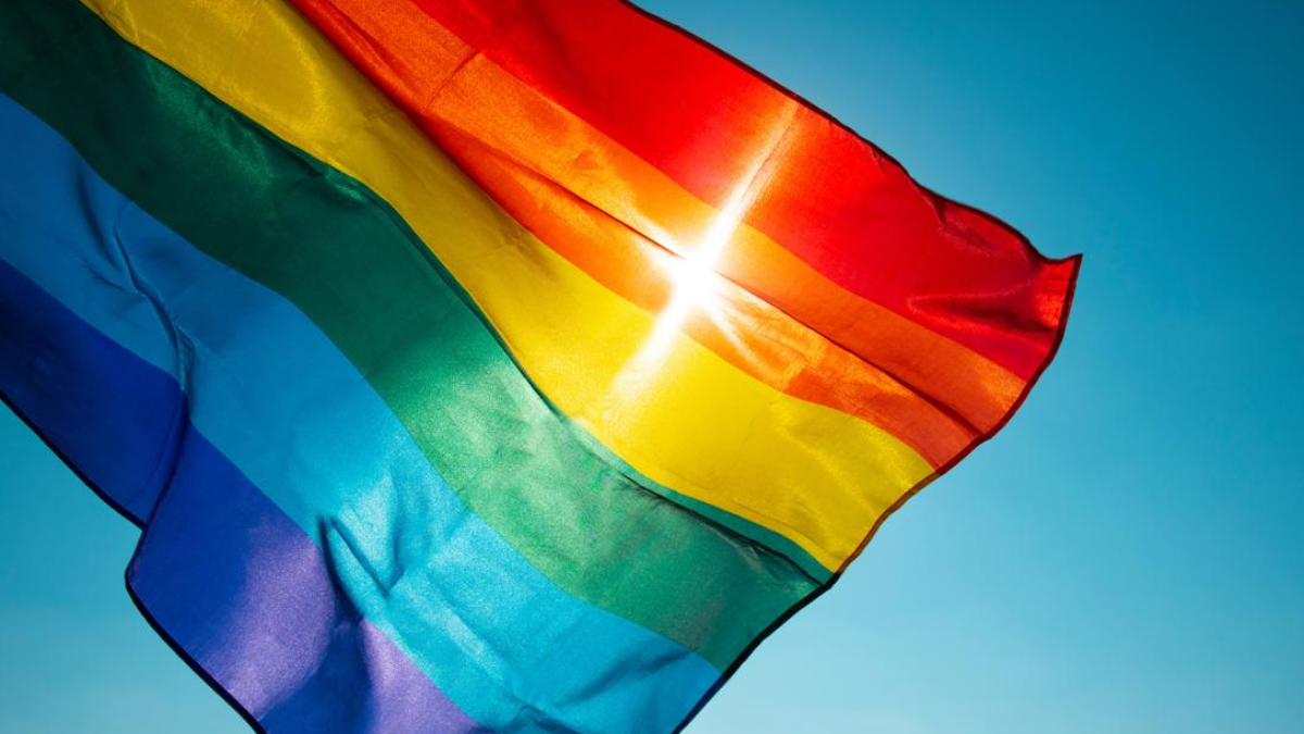 Rainbow Pride flag fluttering in a bright blue sky, sunlight gleaming through the fabric beneath a red stripe.