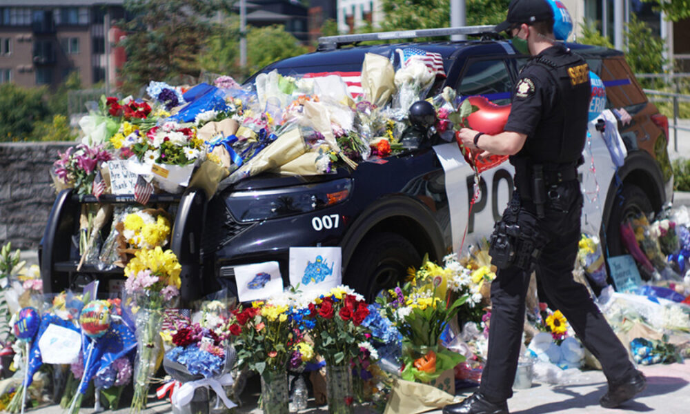 Police SUV covered with flowers, notes, and balloons in a memorial scene; a uniformed officer stands nearby.