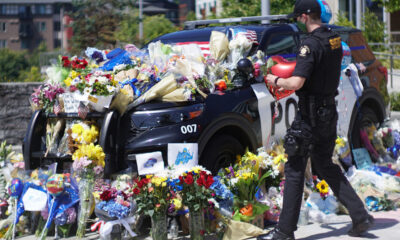 Police SUV covered with flowers, notes, and balloons in a memorial scene; a uniformed officer stands nearby.