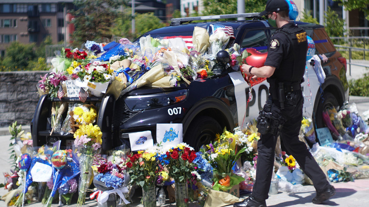 Police SUV covered with flowers, notes, and balloons in a memorial scene; a uniformed officer stands nearby.