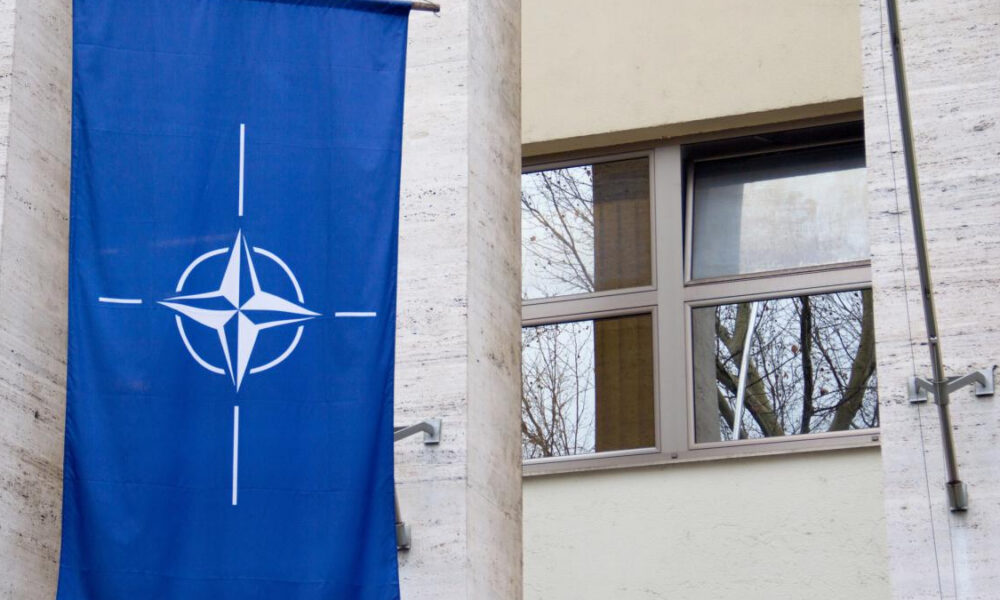 Blue banner with NATO compass emblem hanging on a light-colored stone building beside a window reflecting bare trees in the glass.