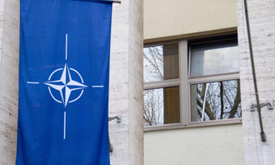 Blue banner with NATO compass emblem hanging on a light-colored stone building beside a window reflecting bare trees in the glass.