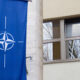 Blue banner with NATO compass emblem hanging on a light-colored stone building beside a window reflecting bare trees in the glass.