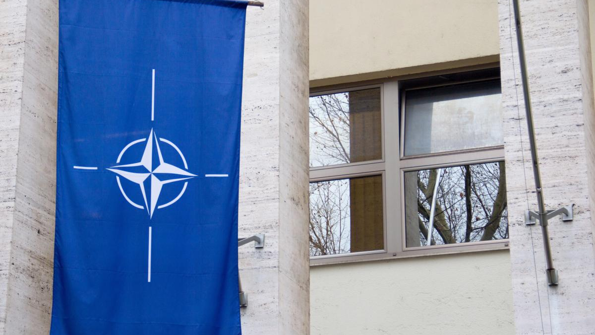 Blue banner with NATO compass emblem hanging on a light-colored stone building beside a window reflecting bare trees in the glass.