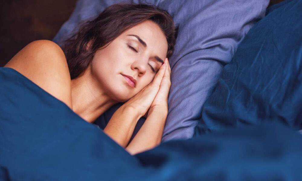 Woman sleeping with hands tucked under her cheek on blue bed linens, eyes closed in a peaceful rest.