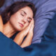 Woman sleeping with hands tucked under her cheek on blue bed linens, eyes closed in a peaceful rest.